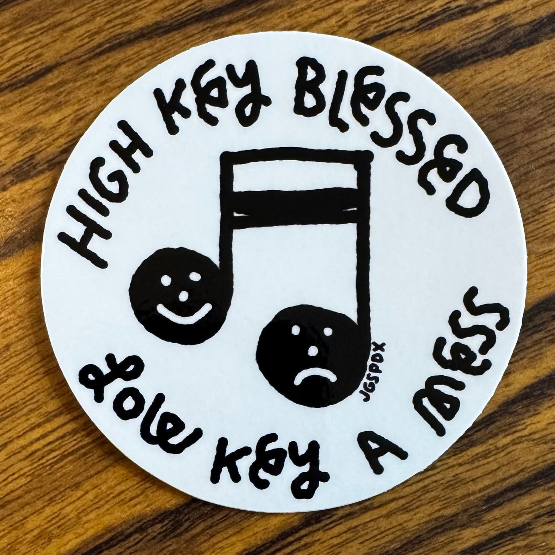 Round sticker with text 'High Key Blessed Low Key A Mess' with a music note in the middle that has a smiley and sad face in the one note.