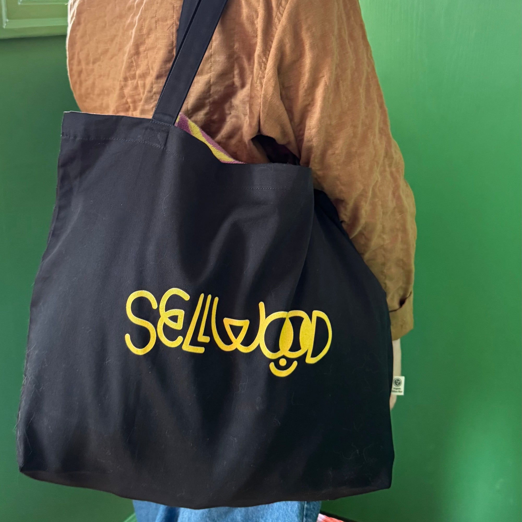 Person holding a black tote bag with 'Sell Out' in yellow text against a green wall.